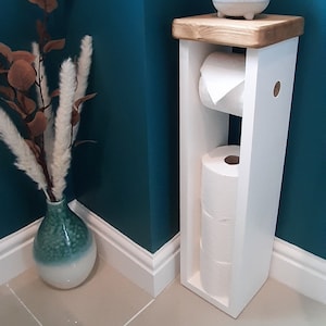 May include: A white wooden toilet paper holder with a natural wood top. The holder contains two rolls of toilet paper. A small potted succulent sits on top. A decorative vase with dried flowers is to the left.