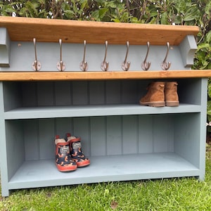 May include: A wooden hallway storage unit with a shelf, hooks, and a top shelf. The unit is painted in a muted blue-grey colour with a natural wood top. Brown boots and red and black wellington boots are on the shelves.