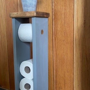 May include: A gray wooden toilet paper holder with three rolls of white toilet paper. A small potted plant sits on top. The holder is painted gray and has a wooden top and a wooden accent on the side.