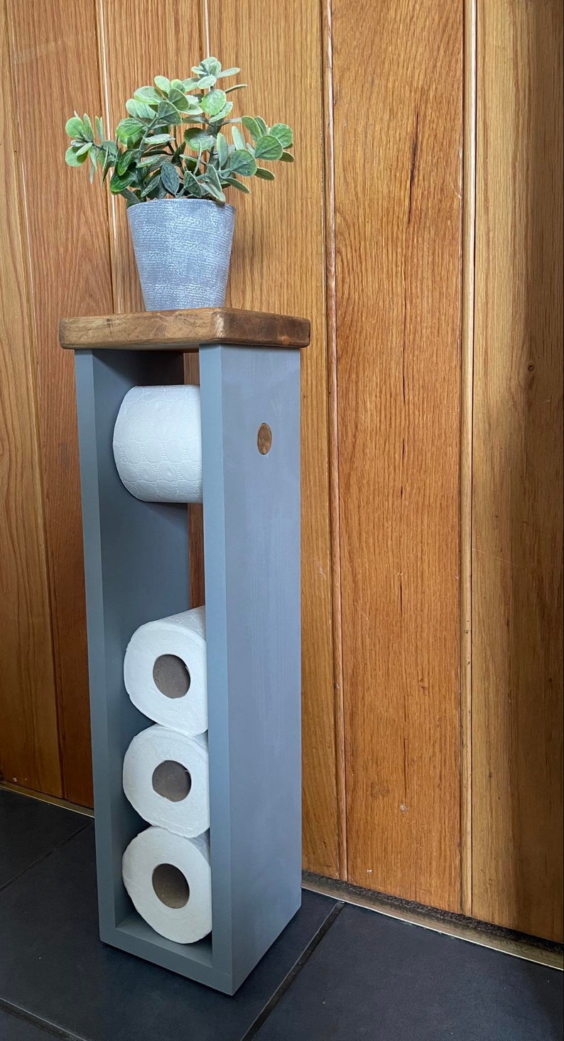 May include: A gray wooden toilet paper holder with three rolls of white toilet paper. A small potted plant sits on top. The holder is painted gray and has a wooden top and a wooden accent on the side.
