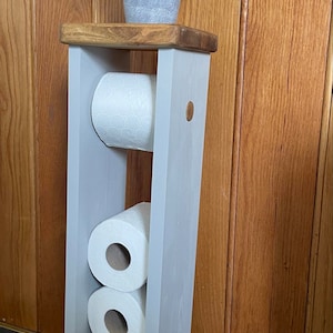 May include: A gray wooden toilet paper holder with a natural wood top, holding three rolls of white toilet paper. A small potted plant with green leaves sits on top. The holder is painted gray and stands against a wooden door.