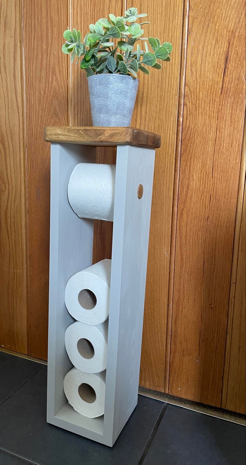 May include: A gray wooden toilet paper holder with a natural wood top, holding three rolls of white toilet paper. A small potted plant with green leaves sits on top. The holder is painted gray and stands against a wooden door.