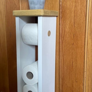 May include: A white wooden toilet paper holder with three rolls of toilet paper. The holder has a natural wood top and a small potted plant. The holder is against a wooden wall.