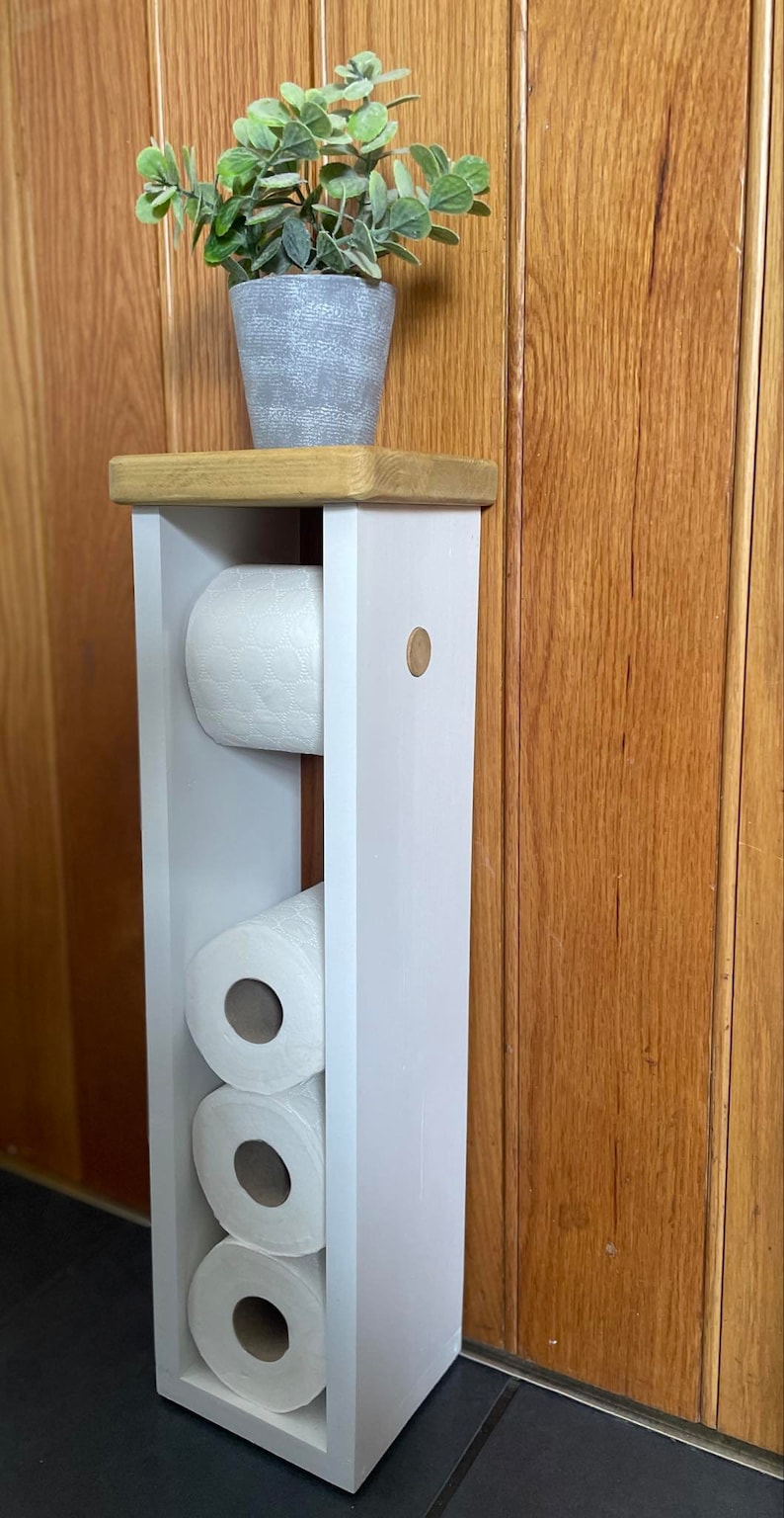 May include: A white wooden toilet paper holder with three rolls of toilet paper. The holder has a natural wood top and a small potted plant. The holder is against a wooden wall.