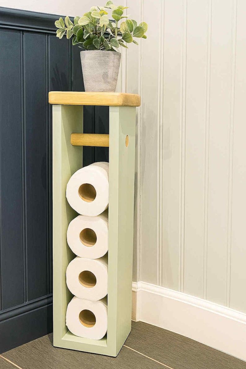 May include: A light green wooden toilet paper holder with a natural wood top and a wooden dowel for holding a roll. The holder contains four rolls of white toilet paper. A potted plant sits on top.
