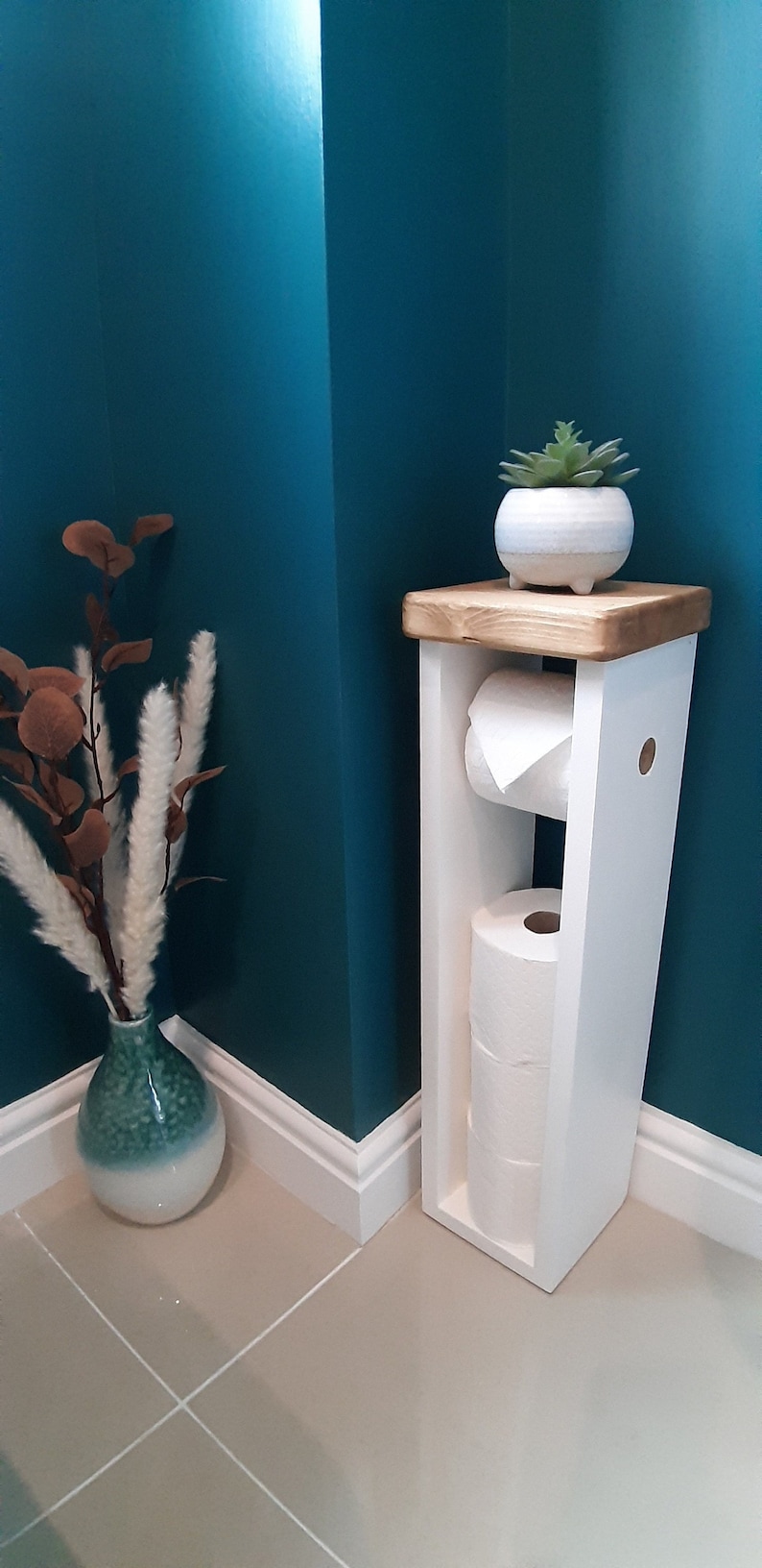 May include: A white wooden toilet paper holder with a natural wood top. The holder contains two rolls of toilet paper. A small potted succulent sits on top. A decorative vase with dried flowers is to the left.