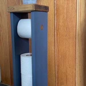 May include: A freestanding, navy blue wooden toilet paper holder with a dark stained top. The holder has two compartments for rolls of toilet paper and a small potted plant on top. The background is a wooden door.