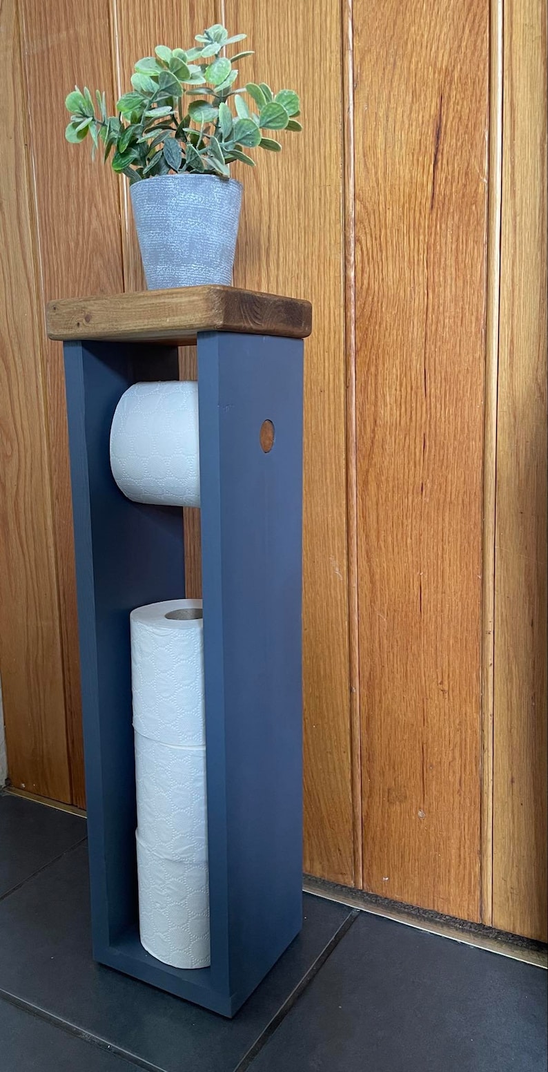 May include: A freestanding, navy blue wooden toilet paper holder with a dark stained top. The holder has two compartments for rolls of toilet paper and a small potted plant on top. The background is a wooden door.