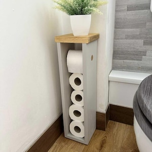 May include: A gray wooden toilet paper holder with a natural wood top, holding several rolls of toilet paper. A white potted plant sits on top. The holder is placed in a bathroom corner, next to a toilet.