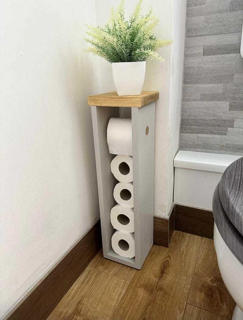 May include: A gray wooden toilet paper holder with a natural wood top, holding several rolls of toilet paper. A white potted plant sits on top. The holder is placed in a bathroom corner, next to a toilet.