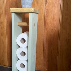 May include: A wooden, freestanding toilet paper holder painted in a light green color. The holder has a top shelf with a small potted plant and holds four rolls of toilet paper. A wooden dowel is present for holding a roll.