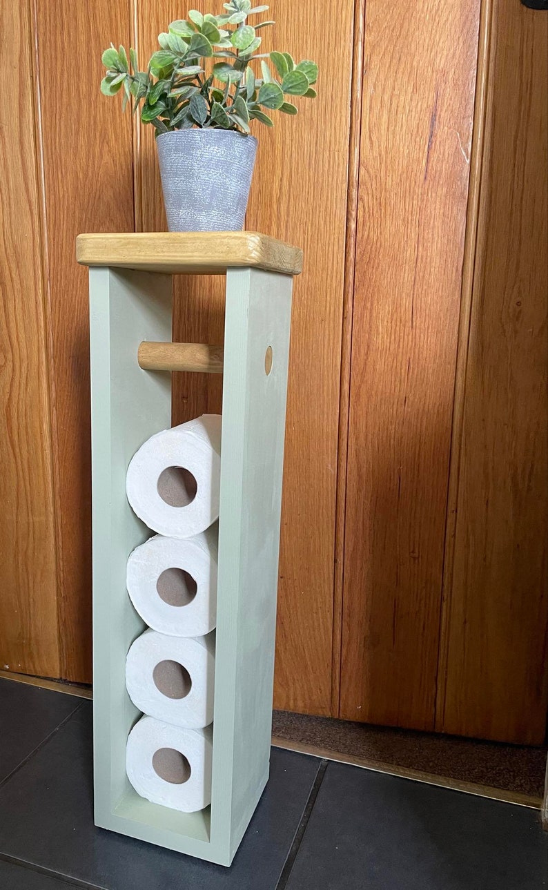 May include: A wooden, freestanding toilet paper holder painted in a light green color. The holder has a top shelf with a small potted plant and holds four rolls of toilet paper. A wooden dowel is present for holding a roll.
