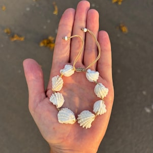 May include: A bracelet featuring seven white, ridged seashells strung together on a tan cord. The bracelet includes an adjustable sliding knot closure with two tan cords ending in small white beads, allowing for a customizable fit on the wrist.
