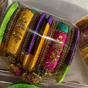 May include: A collection of colorful bangles in a clear plastic package. The bangles are adorned with gold-colored embellishments and feature vibrant hues of yellow, green, purple, and pink. The bangles appear to be made of fabric and are likely for decorative purposes.