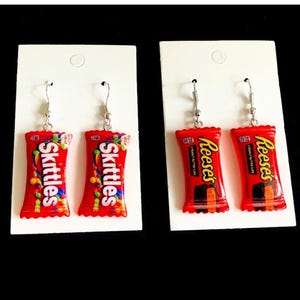 May include: Two pairs of dangle earrings on white display cards. One pair features miniature red Skittles candy packaging replicas, and the other pair features miniature red Reese's peanut butter cup packaging replicas with silver hooks.