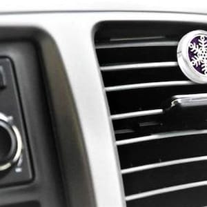 May include: A silver car air freshener with a purple snowflake design. The air freshener is attached to a car's air vent.