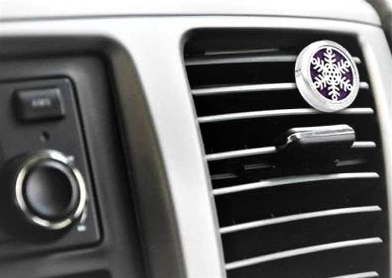 May include: A silver car air freshener with a purple snowflake design. The air freshener is attached to a car's air vent.