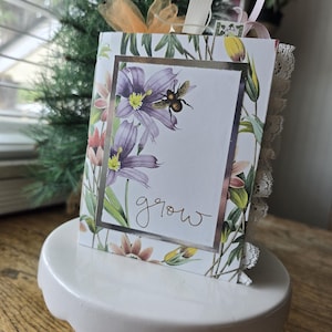 May include: A decorative card with a floral design featuring purple flowers and a bee. The card has the word "grow" in cursive and is trimmed with lace. It is displayed on a white cake stand.