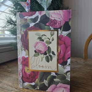 May include: A floral journal with a pink rose design and the word "bloom" in gold script. The journal has a lace trim on the left side and a butterfly and floral ribbon bookmark. The background features a wooden table.
