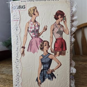May include: A rectangular journal cover featuring a vintage sewing pattern illustration of three figures in summer clothing. The cover is decorated with white lace trim along the edges and includes the text Ask for the Simplicity Pattern Book.