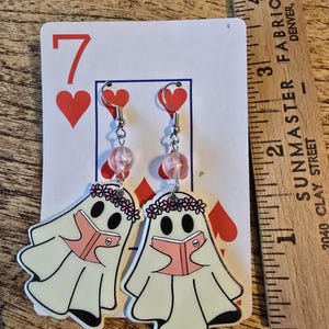 May include: A pair of whimsical earrings featuring ghost figures reading books. The earrings have pink accents, including heart-shaped hooks and beaded dangles. The ghosts are wearing flower crowns. The earrings are displayed on a playing card.