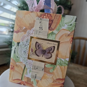 May include: A rectangular greeting card featuring a floral pattern in shades of orange and green. The card is decorated with a white lace doily, a strip of vintage sheet music, and a square sticker depicting a butterfly with the text Player's Cigarette.