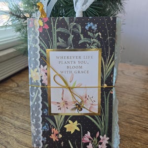 May include: A floral journal with a black background and gold accents. The cover features a botanical design with various flowers and the text "WHEREVER LIFE PLANTS YOU, BLOOM WITH GRACE." A gold string wraps around the journal, and lace trim adorns the edges.