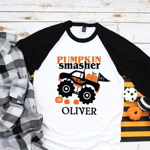 Puede incluir: Camiseta de béisbol de manga larga blanca y negra con un gráfico de camión monstruo negro, el texto "Pumpkin Smasher #1" y el nombre "Oliver".