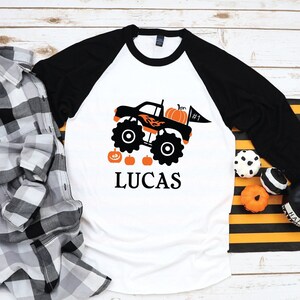 May include: White and black baseball tee shirt with a black monster truck carrying a pumpkin and a flag that says #1. The truck is on a white background with four small pumpkins and the name Lucas in black letters.
