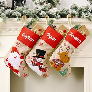Può includere: Tre calze di Natale personalizzate con bordi rossi e bianchi. Le calze sono appese a un camino bianco e hanno nomi ricamati: Sophia, Ryan e Amelia. Ogni calza ha un diverso design festivo: Babbo Natale, un pupazzo di neve e una renna.