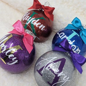 K&ouml;nnte beinhalten: Vier personalisierte Weihnachtskugeln mit Glitzer und Namen. Die Kugeln sind rot, lila, blau und silber mit wei&szlig;en Buchstaben. Die Namen sind Kayden, Ashley, Landon und Avery.