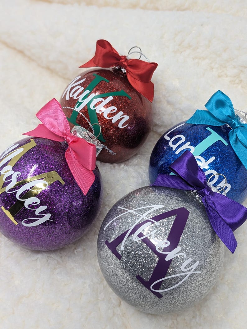 K&ouml;nnte beinhalten: Vier personalisierte Weihnachtskugeln mit Glitzer und Namen. Die Kugeln sind rot, lila, blau und silber mit wei&szlig;en Buchstaben. Die Namen sind Kayden, Ashley, Landon und Avery.