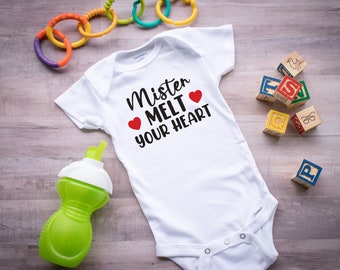 May include: White baby onesie with the text "Mister Melt Your Heart" in black with two red hearts.  The onesie is lying on a wooden surface with a green sippy cup, a colorful toy, and wooden blocks with letters.