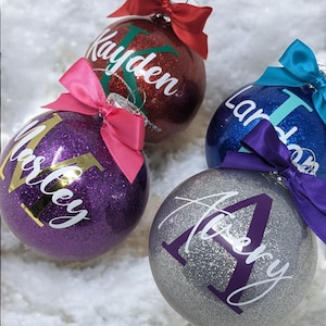 K&ouml;nnte beinhalten: Vier personalisierte Weihnachtskugeln mit Glitzer. Die Kugeln sind rot, lila, blau und silber. Die Namen "Kayden", "Miley", "Laney" und "Avery" sind auf den Kugeln geschrieben.