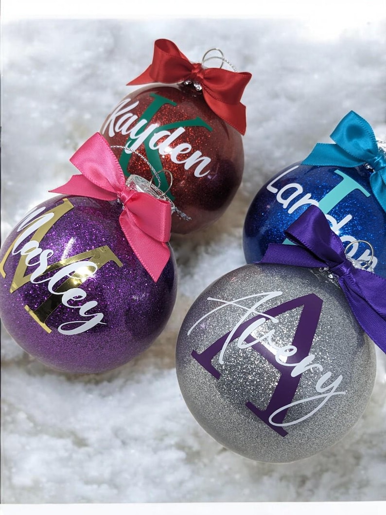 K&ouml;nnte beinhalten: Vier personalisierte Weihnachtskugeln mit Glitzer. Die Kugeln sind rot, lila, blau und silber. Die Namen "Kayden", "Miley", "Laney" und "Avery" sind auf den Kugeln geschrieben.