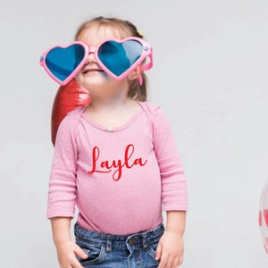 May include: A young child wearing pink heart-shaped sunglasses and a pink long-sleeved bodysuit with the name "Layla" in red script. The child is smiling, with balloons in the background.
