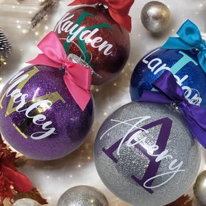 K&ouml;nnte beinhalten: Vier personalisierte Weihnachtskugeln mit Glitzer. Die Kugeln sind rot, lila, blau und silber mit goldenen und wei&szlig;en Buchstaben. Die Namen auf den Kugeln sind Kayden, Mastey, Landon und Avery.