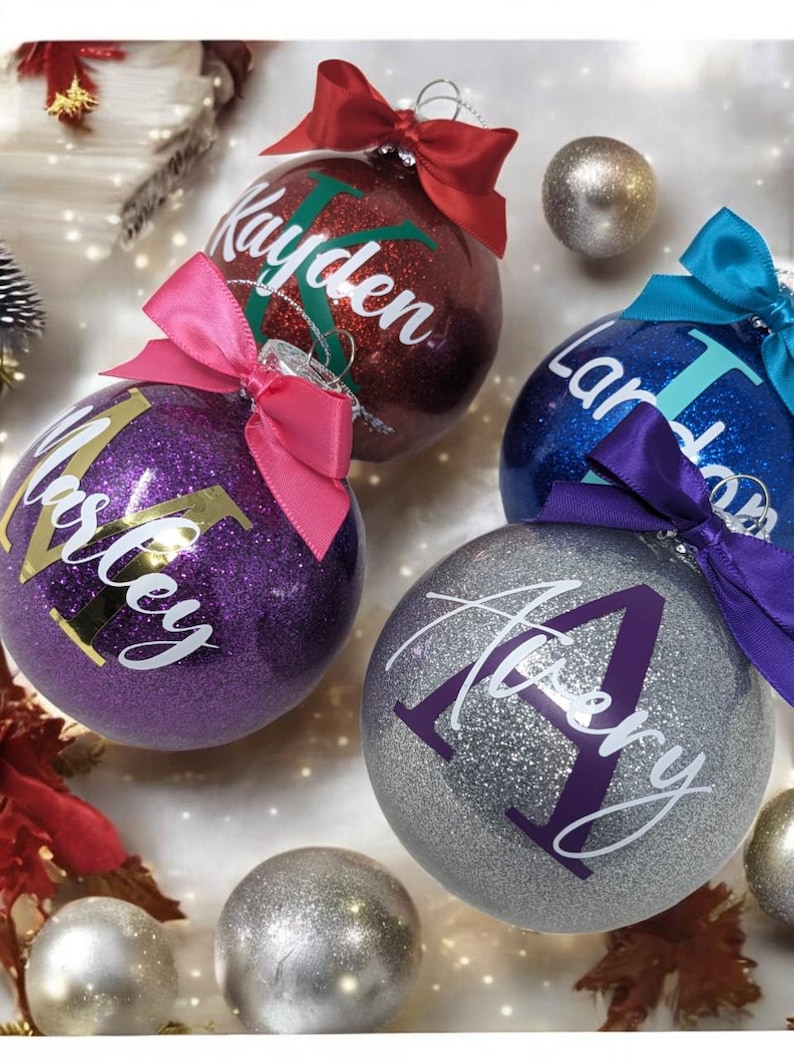 K&ouml;nnte beinhalten: Vier personalisierte Weihnachtskugeln mit Glitzer. Die Kugeln sind rot, lila, blau und silber mit goldenen und wei&szlig;en Buchstaben. Die Namen auf den Kugeln sind Kayden, Mastey, Landon und Avery.