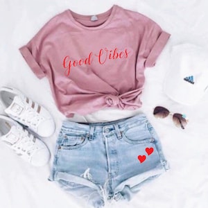 May include: A casual outfit featuring a dusty rose t-shirt with the words "Good Vibes" in red script, paired with light-wash denim shorts adorned with two red heart decals. White sneakers, a white cap, and sunglasses complete the ensemble.