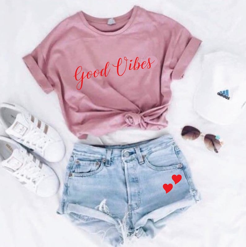 May include: A casual outfit featuring a dusty rose t-shirt with the words "Good Vibes" in red script, paired with light-wash denim shorts adorned with two red heart decals. White sneakers, a white cap, and sunglasses complete the ensemble.