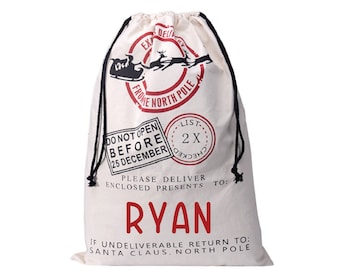 May include: A white canvas Santa sack with red and black details. The sack has a drawstring closure and features a graphic of Santa's sleigh with reindeer, a stamp that says "Do Not Open Before 25 December", and the text "Please Deliver Enclosed Presents To: RYAN. If Undeliverable Return To: Santa Claus, North Pole."
