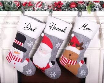 May include: Three gray Christmas stockings with white fur trim. The stockings are decorated with snowflakes and feature a snowman, Santa Claus, and a reindeer. The stockings are personalized with the names "Dad", "Hannah", and "Mom".