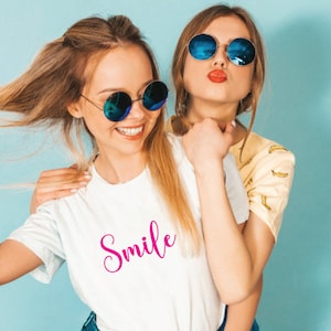 May include: Two young people wearing sunglasses pose for a photo. One wears a white t-shirt with the word "Smile" in pink script. The other wears a yellow shirt with a banana pattern. Both are smiling and looking at the camera.