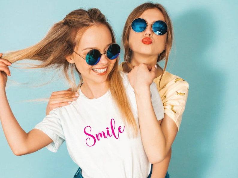 May include: Two young people wearing sunglasses pose for a photo. One wears a white t-shirt with the word "Smile" in pink script. The other wears a yellow shirt with a banana pattern. Both are smiling and looking at the camera.