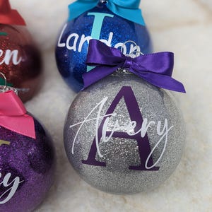 K&ouml;nnte beinhalten: Drei silberne Glitzer-Weihnachtskugeln mit personalisierten Namen. Die Kugeln sind mit violetten, rosa und blauen Schleifen verziert. Die Namen auf den Kugeln sind Landon, Avery und Lily.