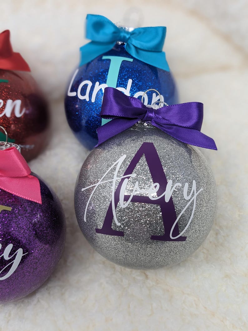 K&ouml;nnte beinhalten: Drei silberne Glitzer-Weihnachtskugeln mit personalisierten Namen. Die Kugeln sind mit violetten, rosa und blauen Schleifen verziert. Die Namen auf den Kugeln sind Landon, Avery und Lily.