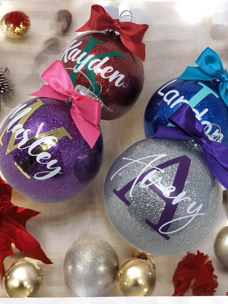 K&ouml;nnte beinhalten: Vier glitzernde Weihnachtskugeln mit darauf geschriebenen Namen. Die Kugeln sind rot, lila, blau und silber. Die Namen auf den Kugeln sind Kayden, Marley, Landon und Avery.