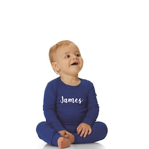 May include: A baby wearing a navy blue pajama set with the name "James" in white script on the chest. The long-sleeved top and pants are a solid color, and the baby is sitting with legs crossed.