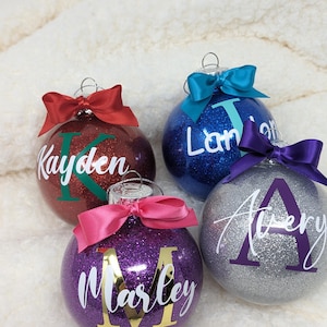 K&ouml;nnte beinhalten: Vier personalisierte Weihnachtskugeln mit Glitzer. Die Kugeln sind rot, blau, lila und silber. Jede Kugel hat einen anderen Namen: Kayden, Landon, Marley und Avery.