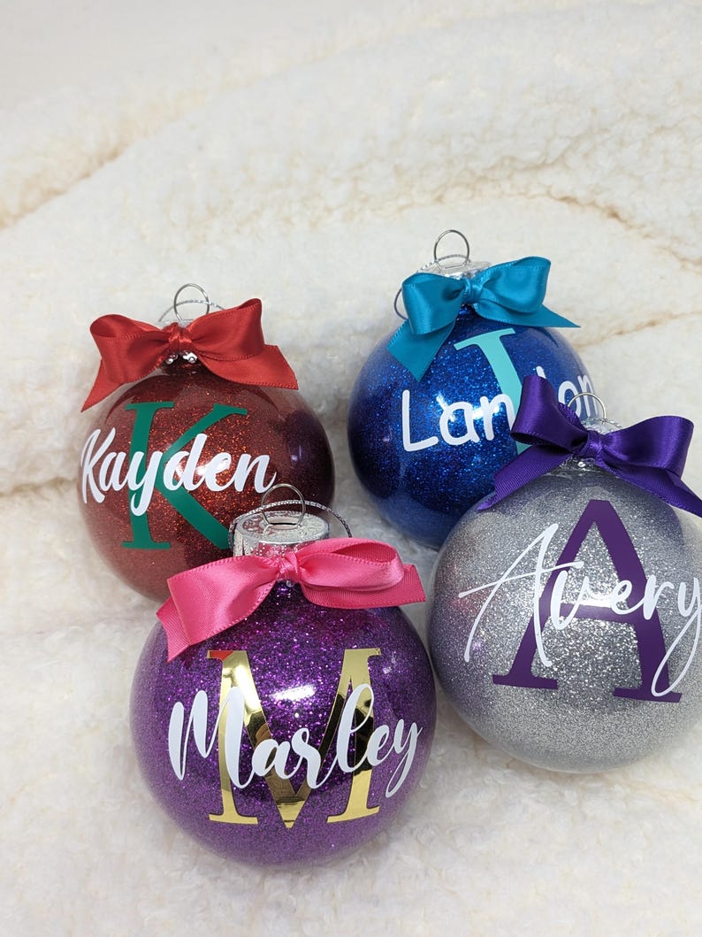K&ouml;nnte beinhalten: Vier personalisierte Weihnachtskugeln mit Glitzer. Die Kugeln sind rot, blau, lila und silber. Jede Kugel hat einen anderen Namen: Kayden, Landon, Marley und Avery.